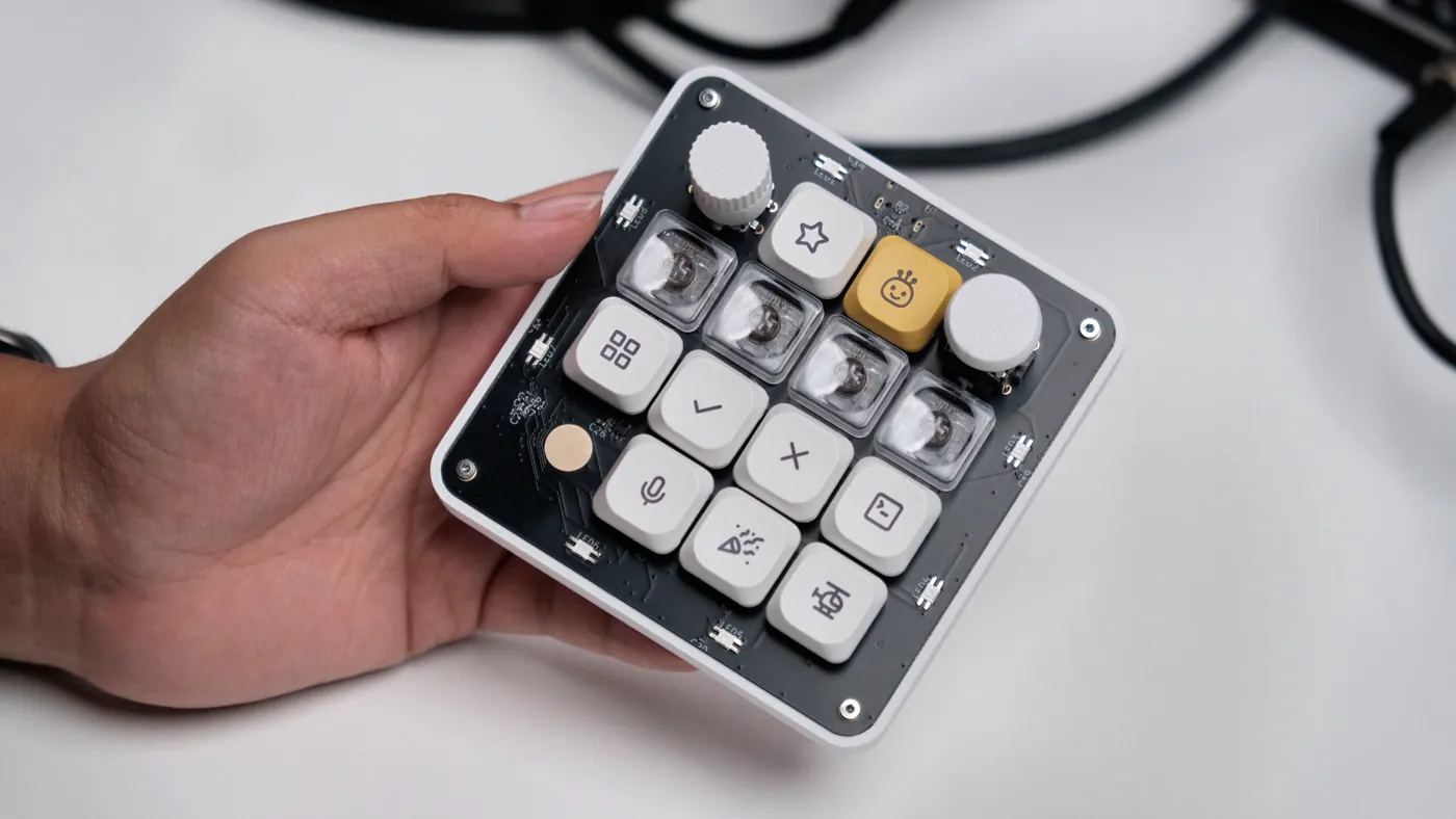 The OpenMicroKbd macropad held in one hand: black PCB in a white printed case, icon keycaps, a knob, a joystick and a yellow accent key